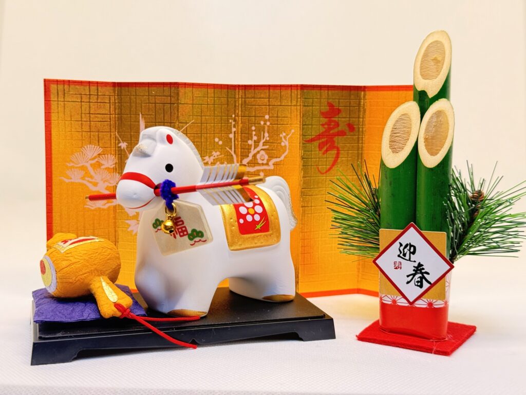 A traditional Japanese New Year decoration featuring a white zodiac horse figurine displayed with a folding gold screen, a wooden mallet, and a bamboo kadomatsu arrangement symbolizing renewal and good fortune.