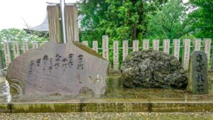 A monument inscribed with the lyrics of Japan’s national anthem “Kimi Ga Yo” stands beside a natural boulder known as “sazare-ishi,” symbolizing the song’s wish for enduring harmony. Photographed at a shrine surrounded by stone pillars and greenery.