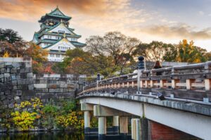 Osaka Castle at sunset with autumn foliage, viewed from a wooden bridge crossing the moat.