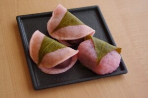 A black plate with two Edo-style sakura mochi (pink crepe-like cakes folded over sweet bean paste) and one Kansai-style domyoji mochi (pink rice cake wrapped in a salted cherry leaf).