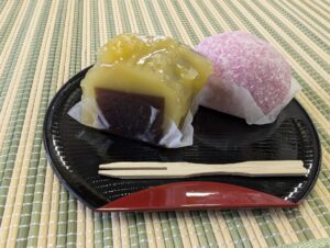 Traditional Japanese sweets (wagashi) served on a black lacquer plate: a bright yellow chestnut yokan (sweet bean jelly) and a pink daifuku rice cake, symbolizing seasonal delicacy and harmony.