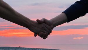 Two people shaking hands against the backdrop of a vivid red and orange sunset over the sea, symbolizing trust, unity, and hope.