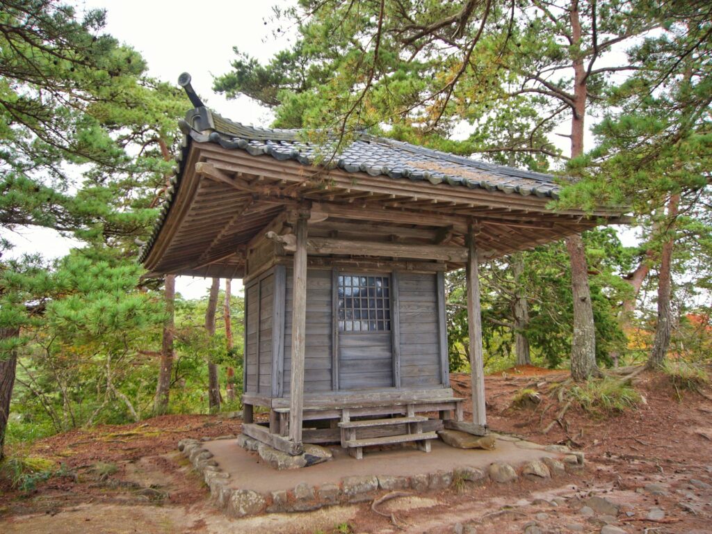 A rustic wooden roadside shrine with a tiled roof, set on a hillside among pine trees—an enduring symbol of local faith and memory in rural Japan.