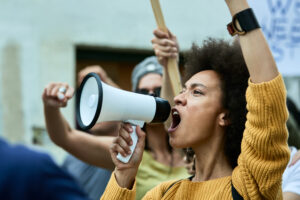A protester passionately shouting through a megaphone, demanding justice as others hold protest signs behind her.