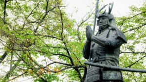 Bronze statue of samurai Yukimori Yamanaka in prayer, wearing armor with antler-shaped horns and a crescent moon crest, set against green spring foliage.
