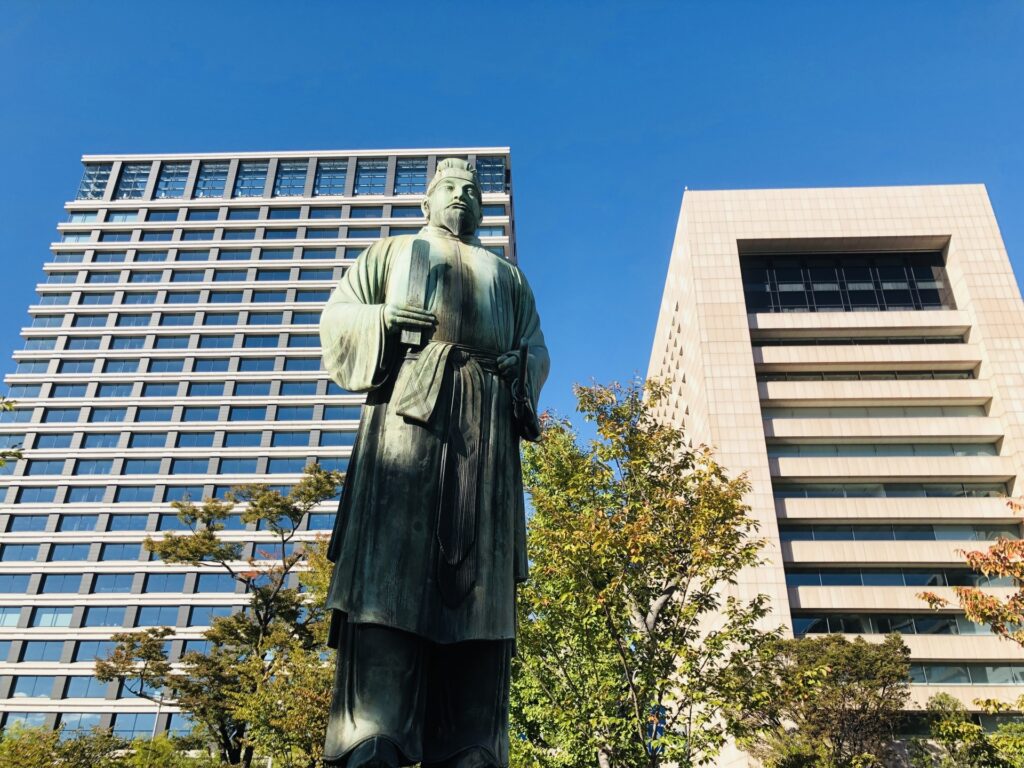 Statue of Wake no Kiyomaro in front of the Imperial Palace, with modern office buildings rising behind. The image symbolizes the connection between ancient loyalty and Japan’s present-day identity.