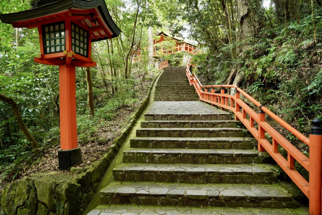 A moss-covered stone stairway lined with red railings and lanterns leading up to Usa Hachiman Shrine in Kyushu, Japan. The path symbolizes centuries of pilgrimage and devotion to the guardian of Japan’s imperial line.