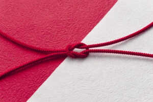 A close-up of a red decorative cord tied in a knot on textured red and white Japanese paper, symbolizing Musubi—the act of binding lives and communities together in harmony.