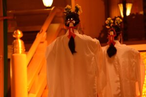 Two Shinto shrine maidens seen from behind, wearing white ceremonial robes and traditional hair ornaments, standing in a warmly lit shrine during a ritual.