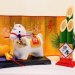 A traditional Japanese New Year decoration featuring a white zodiac horse figurine displayed with a folding gold screen, a wooden mallet, and a bamboo kadomatsu arrangement symbolizing renewal and good fortune.